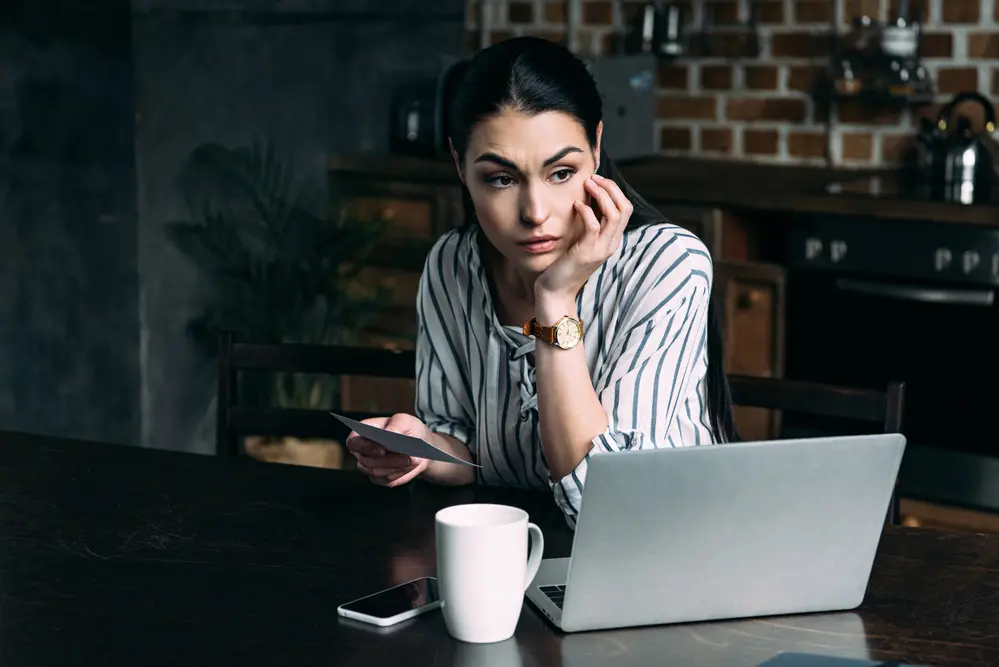 Depressed young woman with smartphone and laptop sitting in the kitchen
