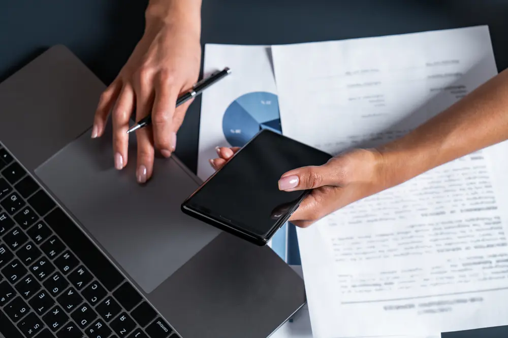 person cross checking information with documents, their phone and their laptop