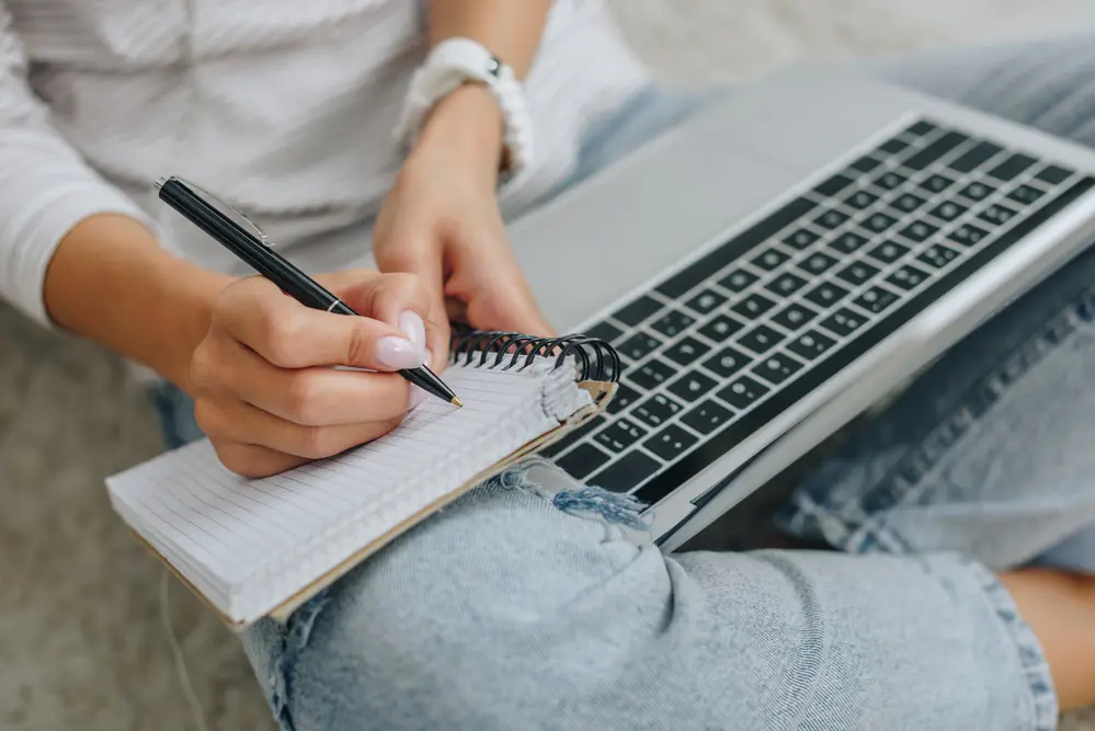 woman writing notes while researching on her laptop